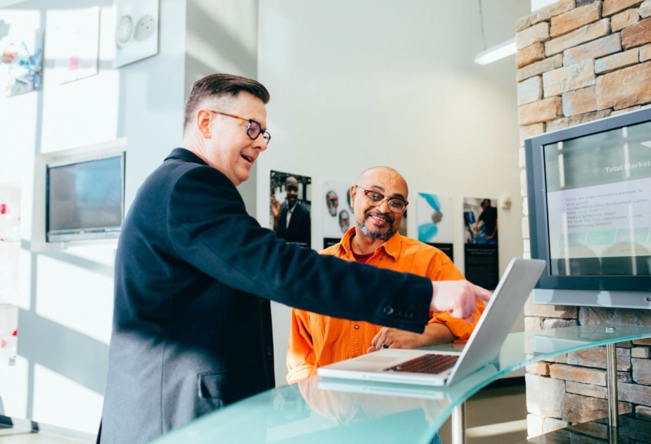 Two men stand together looking at a laptop inside of a business; 0% APR car financing is an attractive option for shoppers