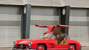 Red 1954 Mercedes-Benz "Gullwing" coupe parked in front of an old Art Deco building with its doors up.