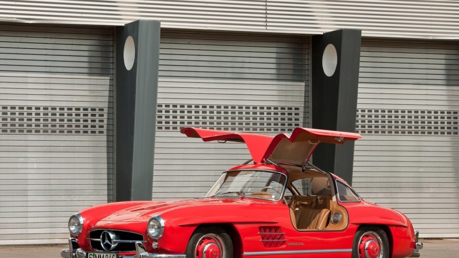 Red 1954 Mercedes-Benz "Gullwing" coupe parked in front of an old Art Deco building with its doors up.