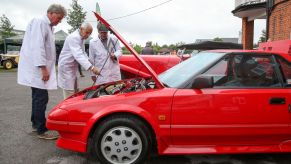A group of Vintage Sports Car Club (VSSC) judges inspecting under the hood of a red 1998 Toyota MR2 Mk1