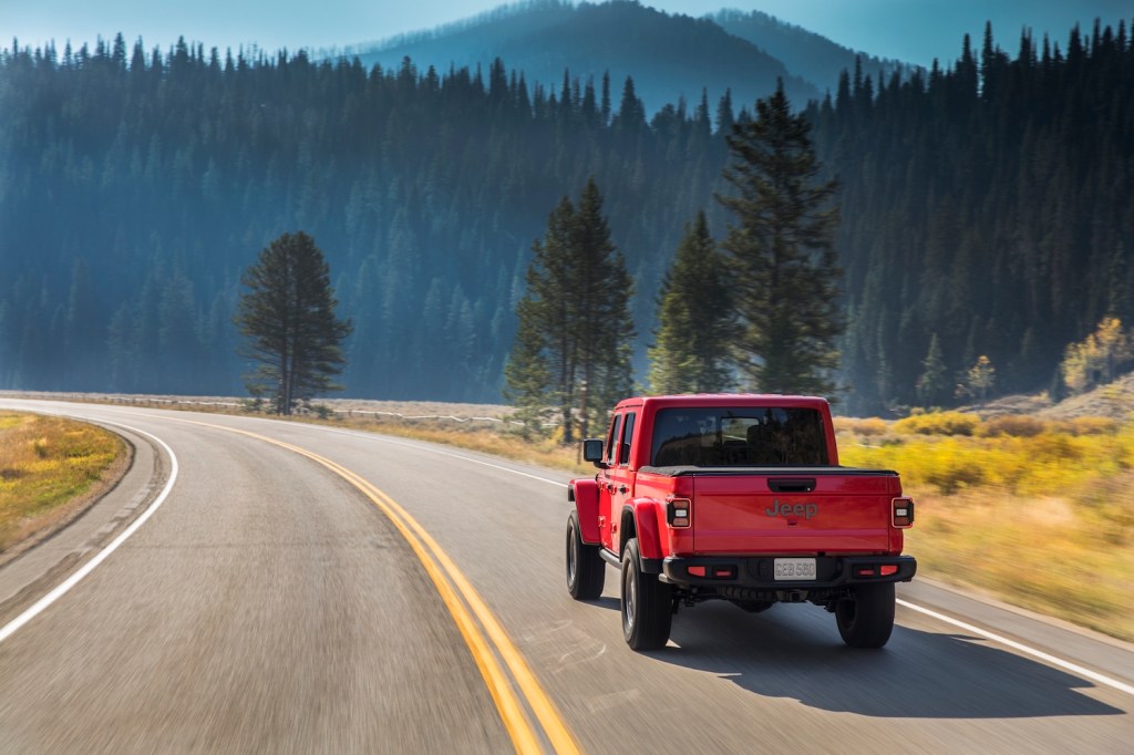 Red Jeep Gladiator pickup truck driving down a paved road with mountains visible in the background.