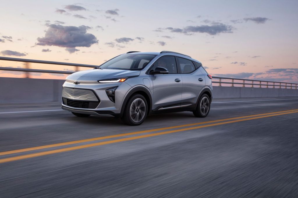 A silver 2022 Chevrolet Bolt EUV driving down a road with a partly cloudy sky in the background. 
