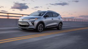 A silver 2022 Chevrolet Bolt EUV driving down a road with a partly cloudy sky in the background.