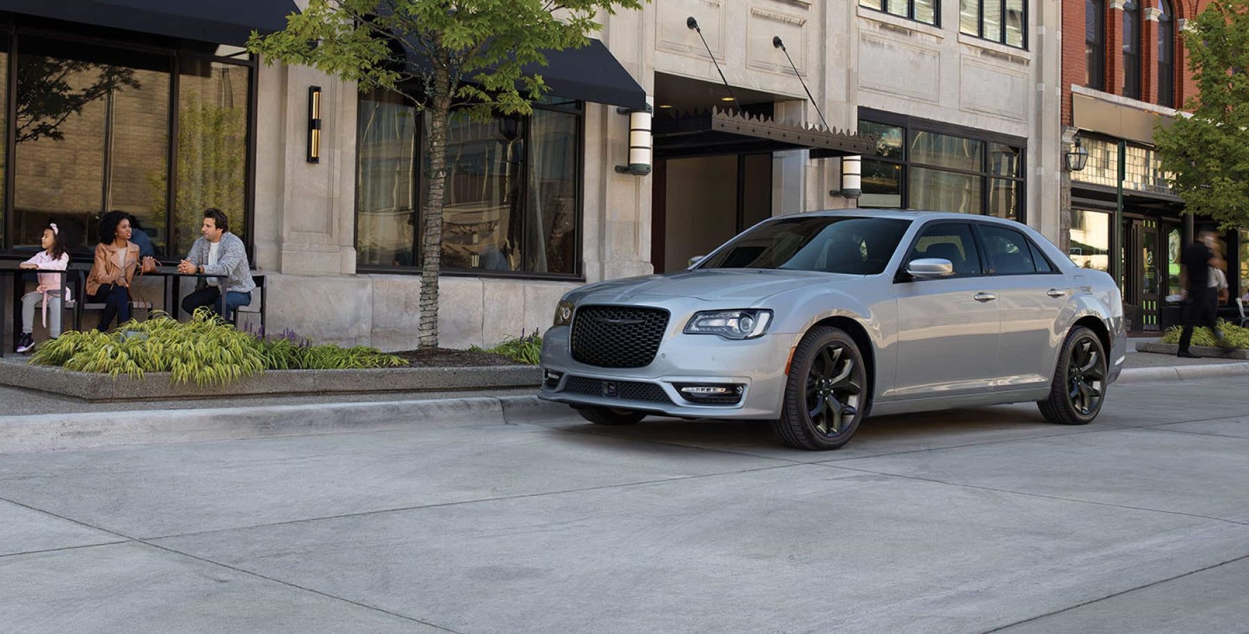 A 2022 Chrysler 300 with a light gray paint color parked on a concrete road near a shopping street and cafe