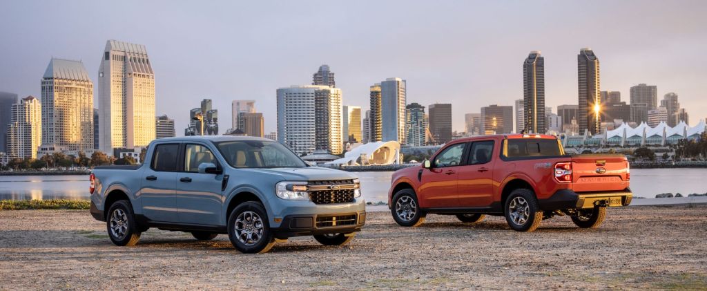 Blue and red/orange 2022 Ford Maverick compact pickup truck models parked on a beach near a waterfront and city skyline