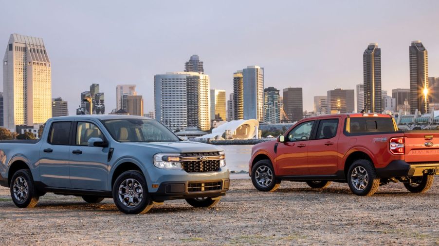 Blue and red/orange 2022 Ford Maverick compact pickup truck models parked on a beach near a waterfront and city skyline