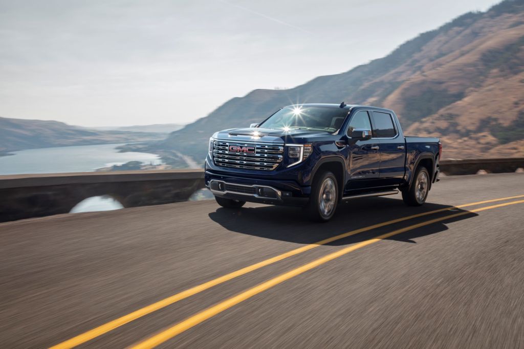 A blue 2022 GMC Sierra on a road in front of a mountain and body of water. 