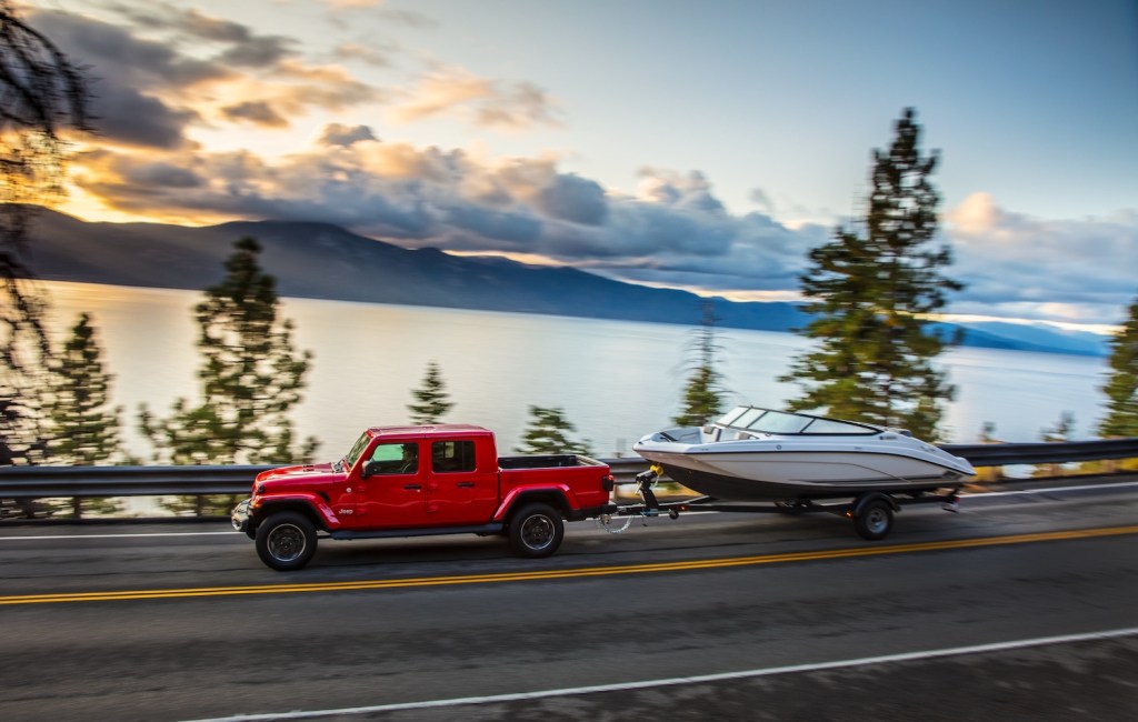 Red Jeep Gladiator pickup truck towing a boat with a lake and mountains visible in the background.