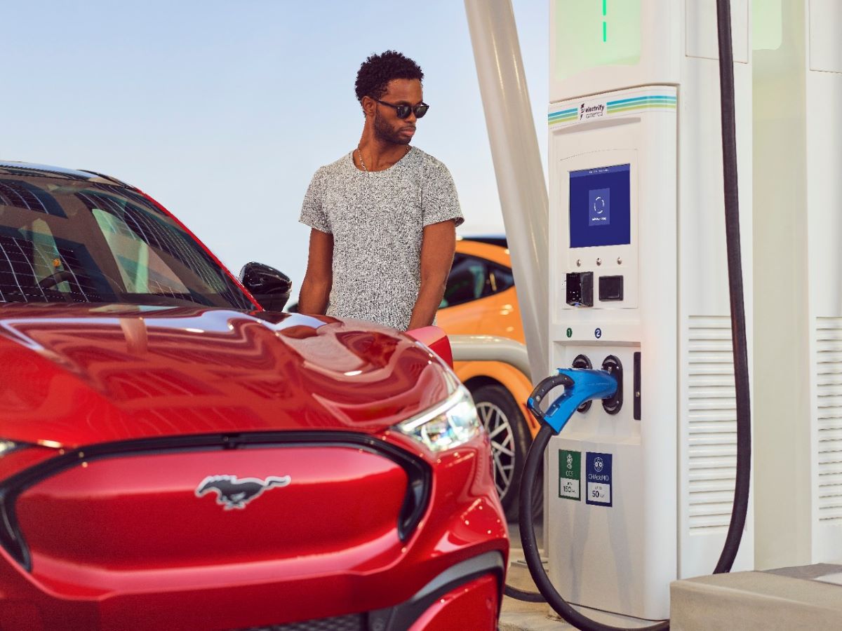 A black man stands next to a red Mustang Mach-E, plugging the electric Mustang into an EV charging station