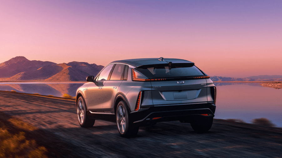 A rearview shot of a gray 2023 Cadillac Lyriq luxury electric SUV model driving near a lake at sunset