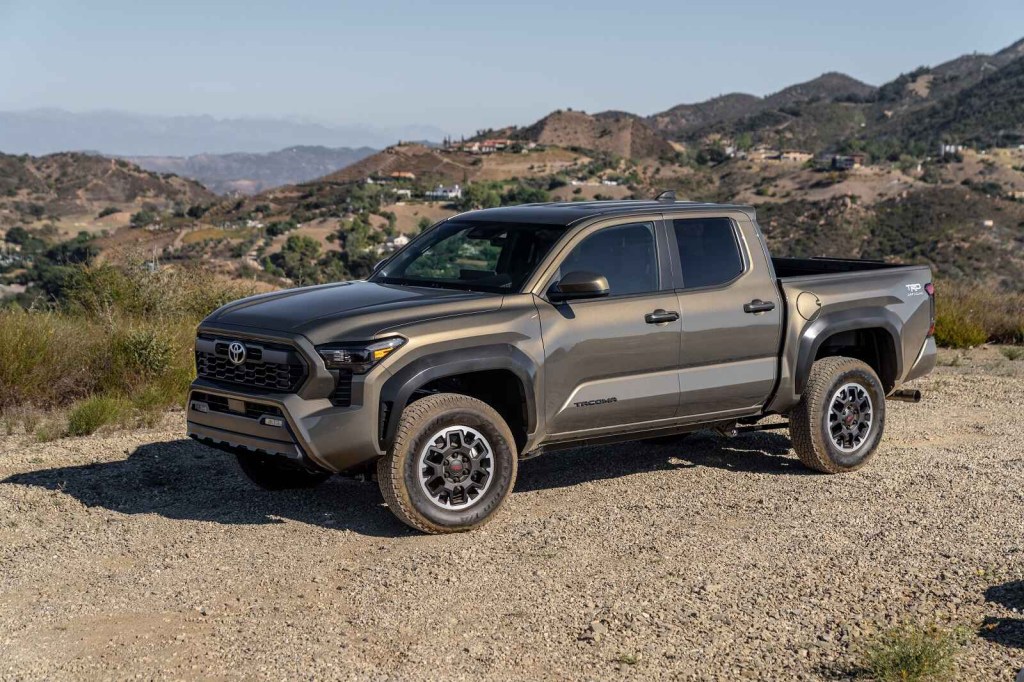 A dark metallic 2024 Toyota Tacoma TRD Off-Road is parked in left front profile view on dirt with rocky hills in the background