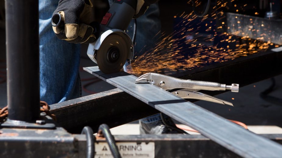 Sparks fly into the air as a worker using an angle grinder to cut a piece of flat stock.