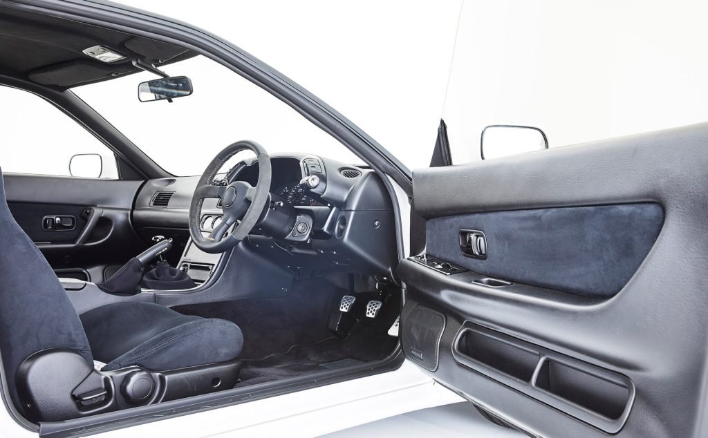 The Alcantara-trimmed front seats and black dashboard of a white Built by Legends Mine's R32 Nissan Skyline GT-R seen through the open driver's door