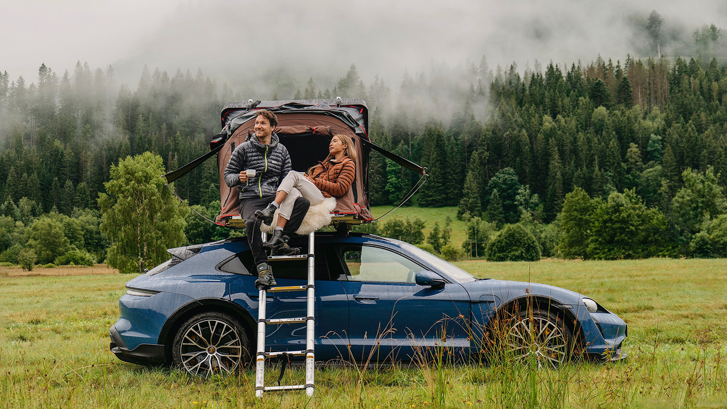 Couple sitting atop blue Porsche roof tent Taycan Turbo S Cross Turismo camping in German black forest