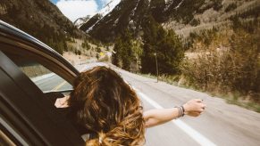 Woman hangs out the passenger window of a car navigating a mountain road.