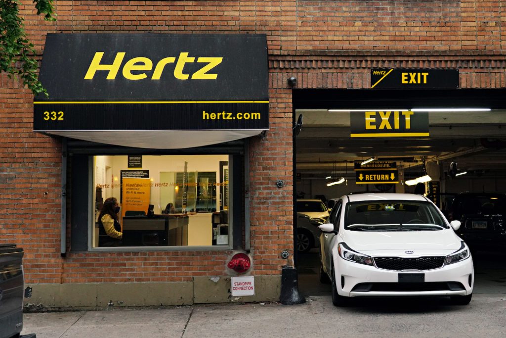 A rental car parked next to the office window of a brick-walled Hertz rental branch.