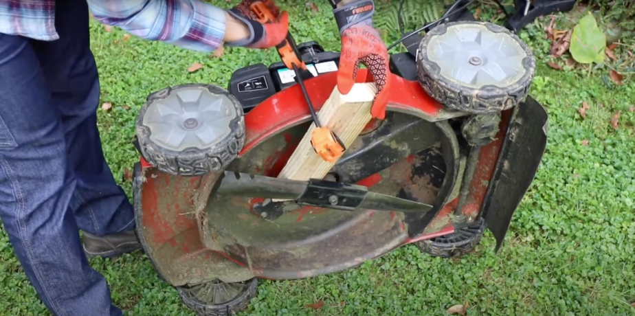 Mechanic clamping a board to the deck of a push lawn mower to hold the blade in place for removal.