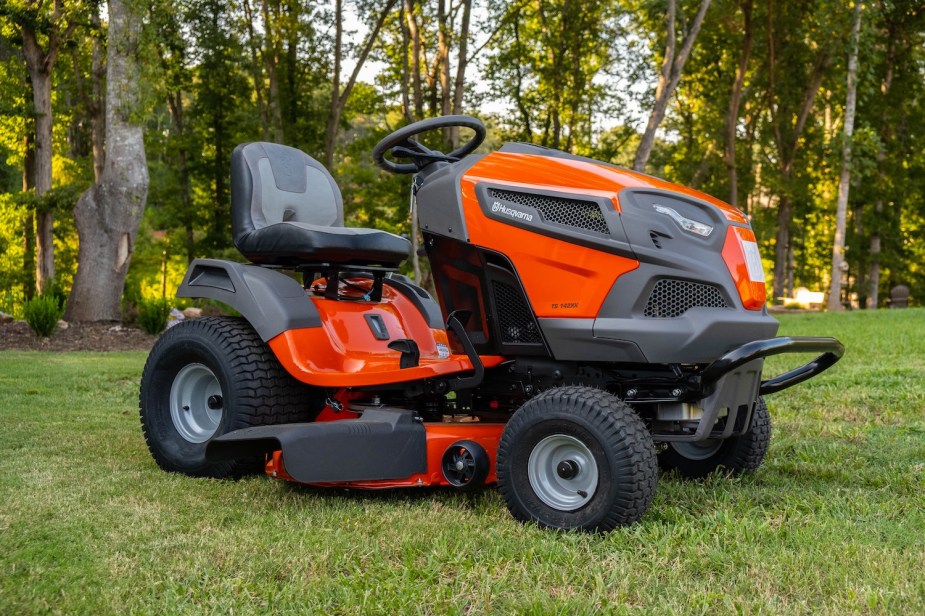 Orange Husqvarna lawn tractor parked on cut grass, by a treeline.