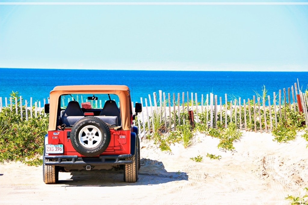 Red soft top Jeep Wrangler parked on the beach, facing the ocean.