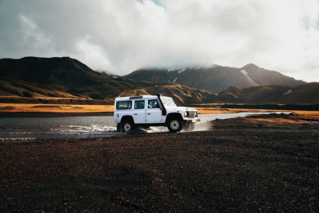 White Land Rover Defender fording a river in front of a misty mountain range.