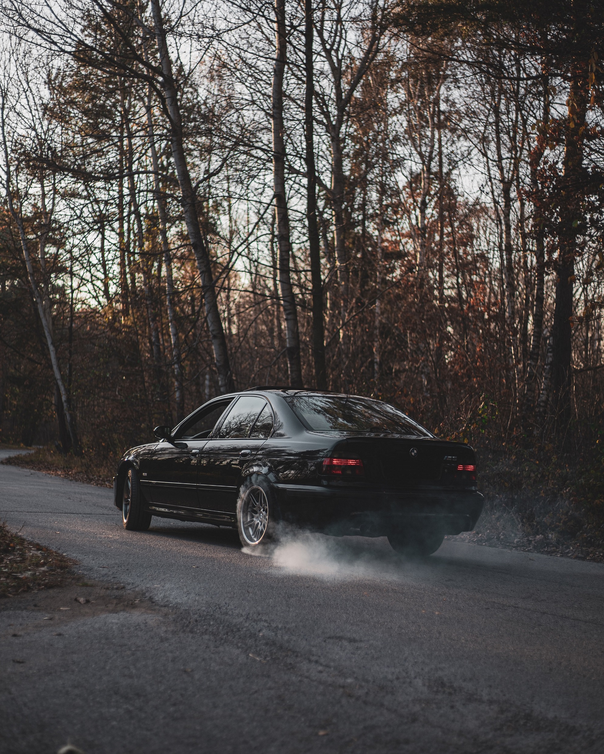 The rear 3/4 view of a black post-facelift BMW E39 M5 doing a burnout on a forest road