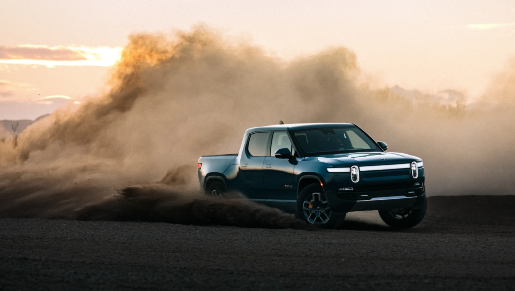 The Rivian R1T all-electric pickup truck model driving at dusk and kicking up dust clouds with its tires