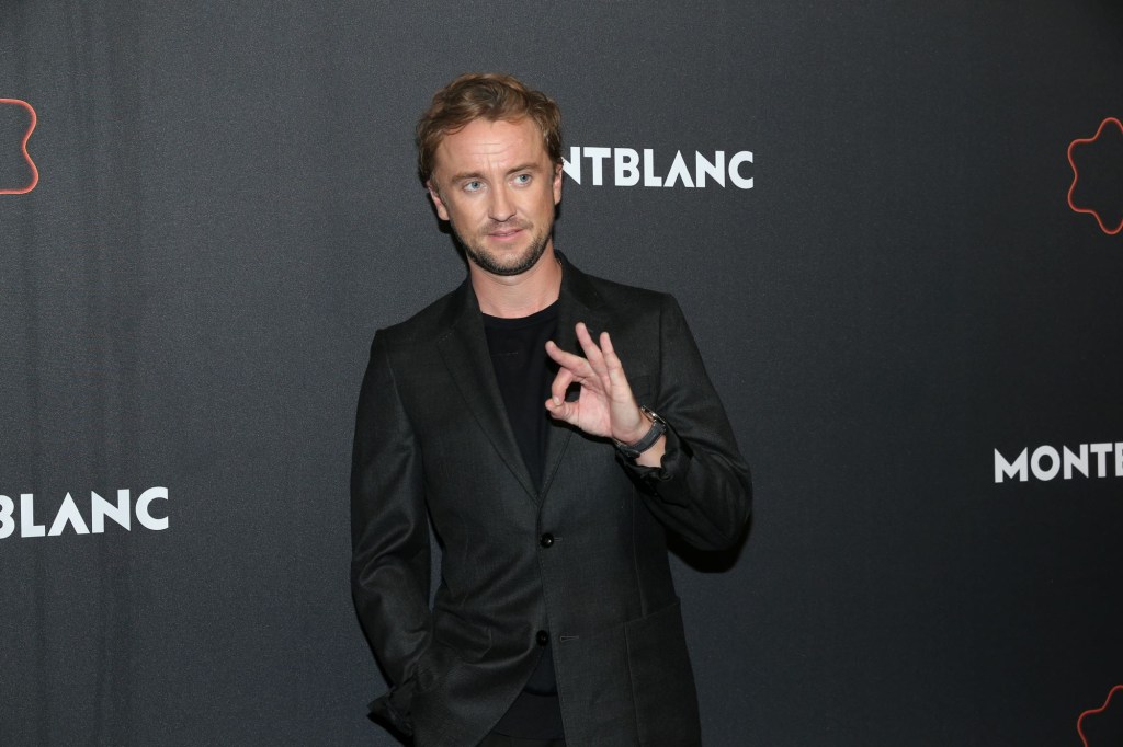 Tom Felton wearing a black suit with a black background. 