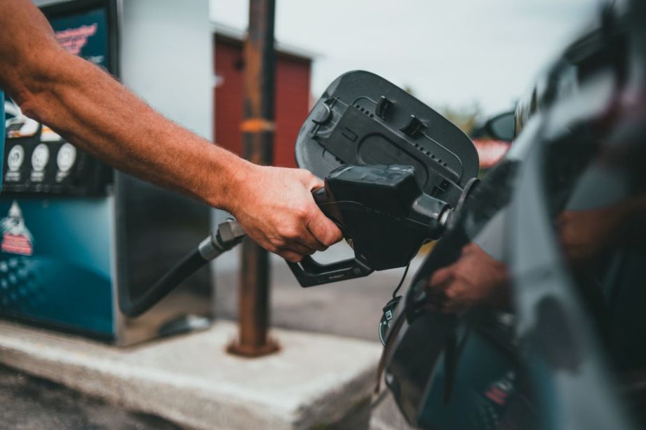 Muscular arm holding a gas pump nozzle up to the fuel cap of a car at a gas station. The best fuel rewards programs of 2022 can help you save money.