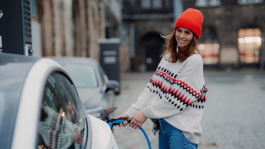 A woman wearing a white sweater and a red hat plugs a charging cord into her reliable used electric car
