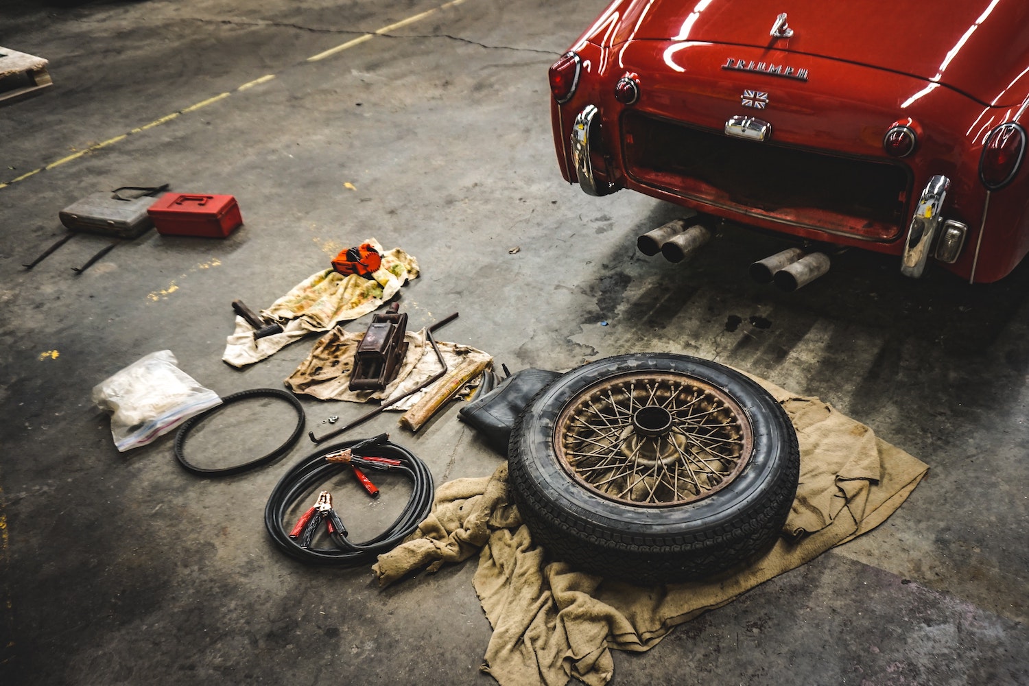 A pile of tools and a spare tire on the floor of a garage behind a Triumph sports car.