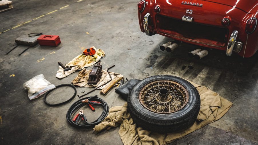 A pile of tools and a spare tire on the floor of a garage behind a Triumph sports car.