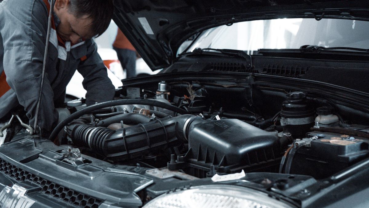 A mechanic leans over an open engine bay, inspecting the vehicle. Routing inspections are important for preventing car fires.