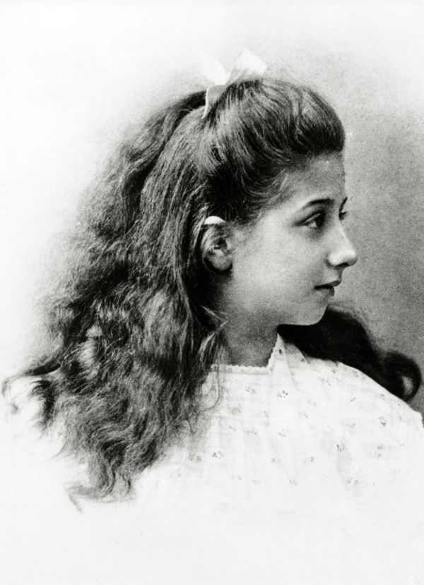 A black-and-white portrait of Mercedes Jellinek as a young girl. She has long, curly hair tied up in a bow