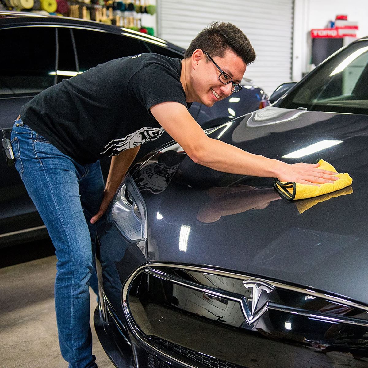 A man wearing jeans, a black t-shirt, and glasses leans over a car with one of the best auto detailing accessories: the simple microfiber towel