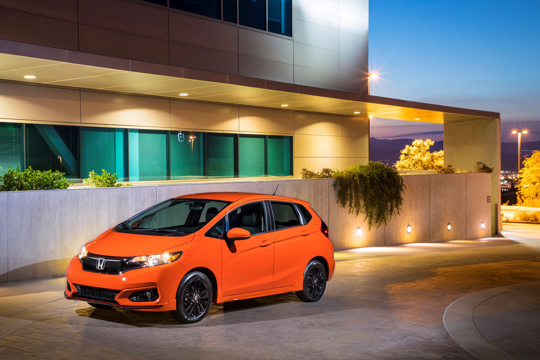 An orange 2018 Honda Fit subcompact hatchback model parked on a stone driveway outside an office building at sunset