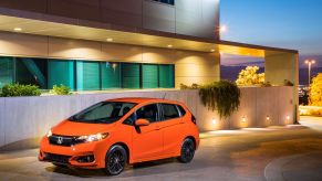 An orange 2018 Honda Fit subcompact hatchback model parked on a stone driveway outside an office building at sunset