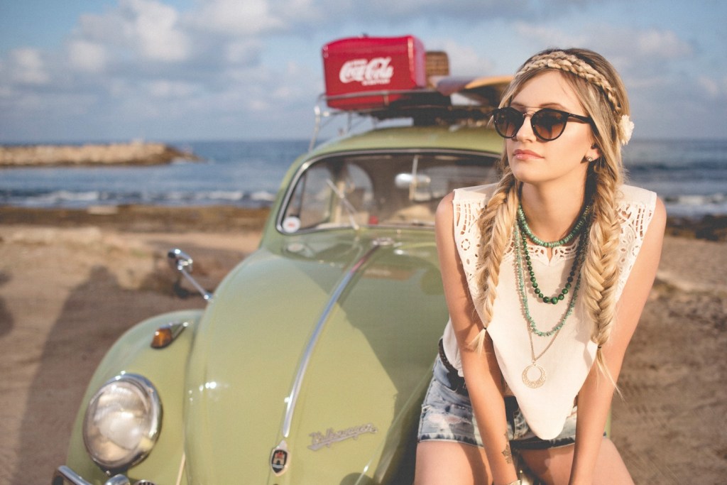 Woman sits on the hood of a green Volkswagen Beetle parked on the beach.