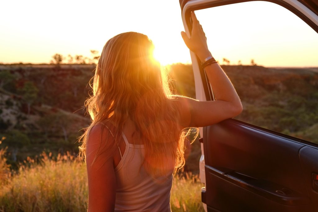 A woman leans against the open door of a parked car, facing the sunset.