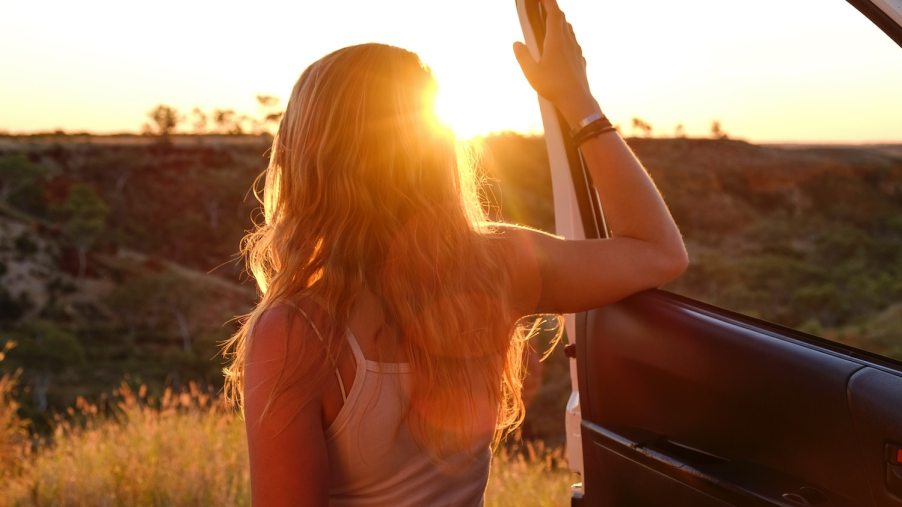 A woman leans against the open door of a parked car, facing the sunset.