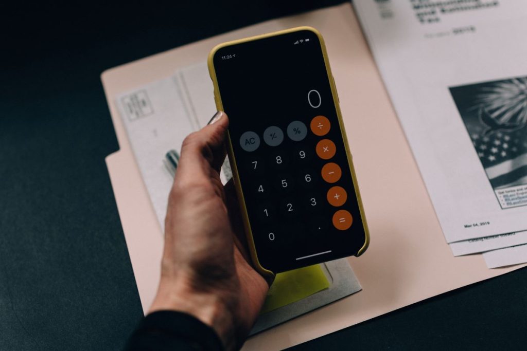 A hand holds a black smartphone in a golden case over a file folder of documents. The calculator app is up on the screen.Are free maintenance plans worth the higher cost of vehicles?