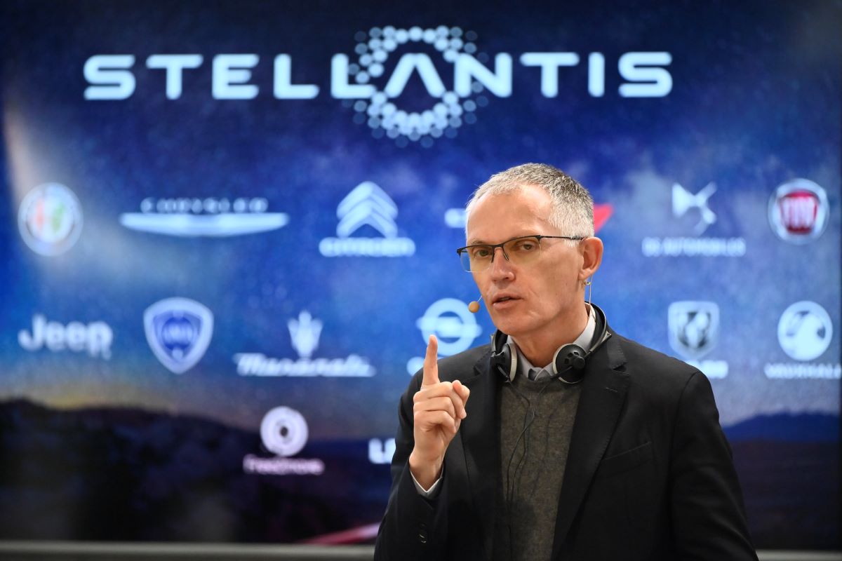 Carlos Tavares, CEO of Stellantis, looks directly into a camera. He holds one finger up while making a point. He's standing in front of a Stallantis banner. 