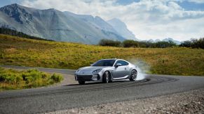 Silver 2022 Subaru BRZ two-door sports car drifting on a curved road with gorgeous mountains in the backdrop. This model is a big step in the Subaru BRZ's history