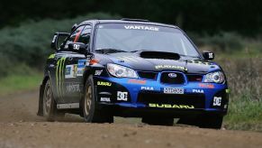 A blue Subaru WRX rally car, covered in sponsorship stickers, races along a dirt track. The Subaru WRX started as a homologation car.