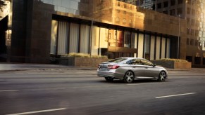 A silver 2021 Honda Accord Hybrid drives down a multi-lane road with a short commercial building in the background
