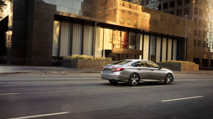 A silver 2021 Honda Accord Hybrid drives down a multi-lane road with a short commercial building in the background