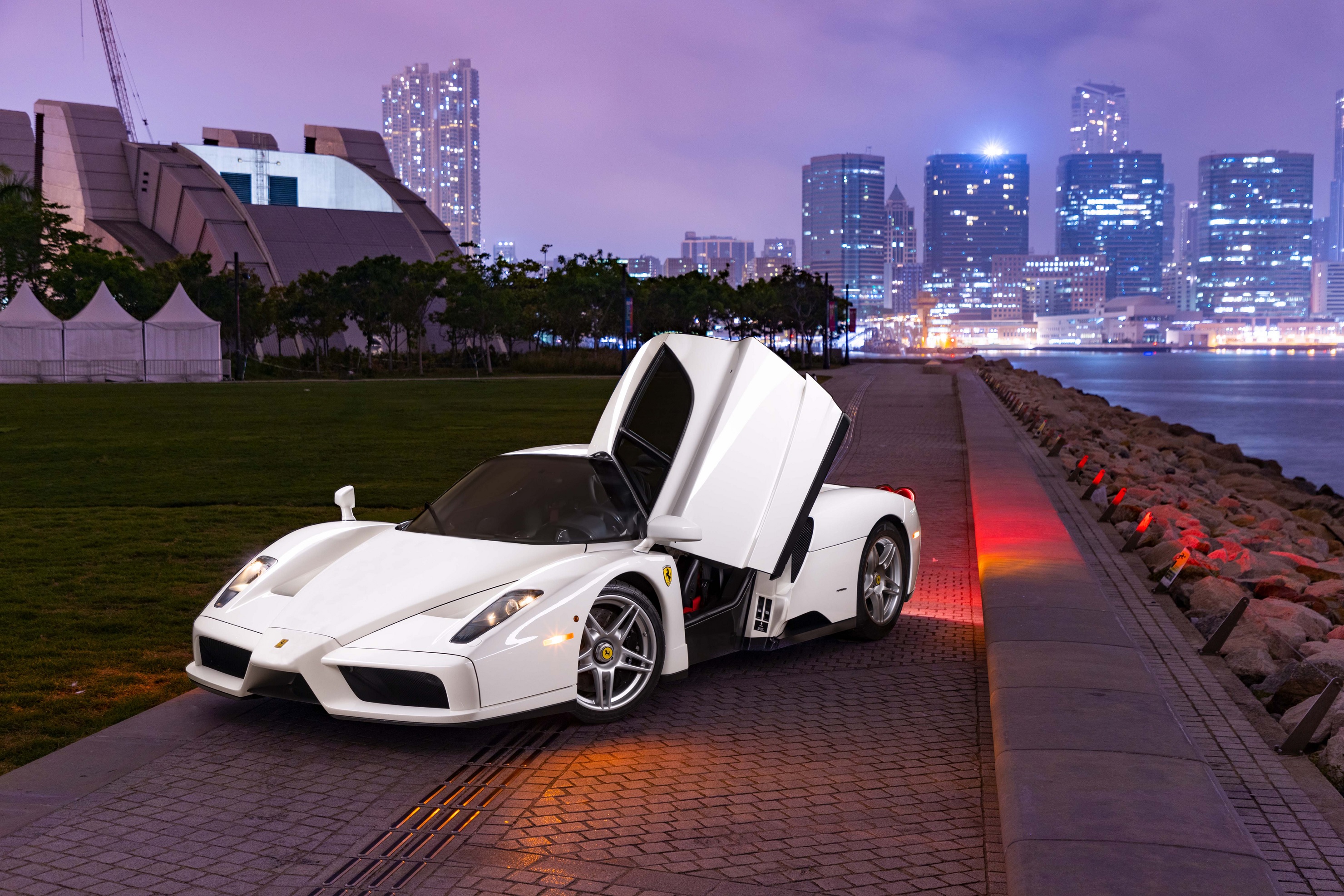 The front 3/4 view of the 1-of-1 Bianco Avus white 2003 Ferrari Enzo with its door open in front of a nighttime city skyline