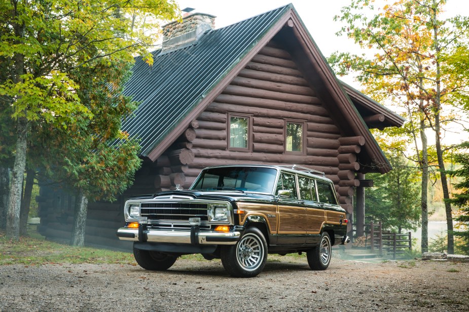 Classic Jeep Grand Wagoneer SUV parked in front of a log cabin, a lake and trees visible in the background.