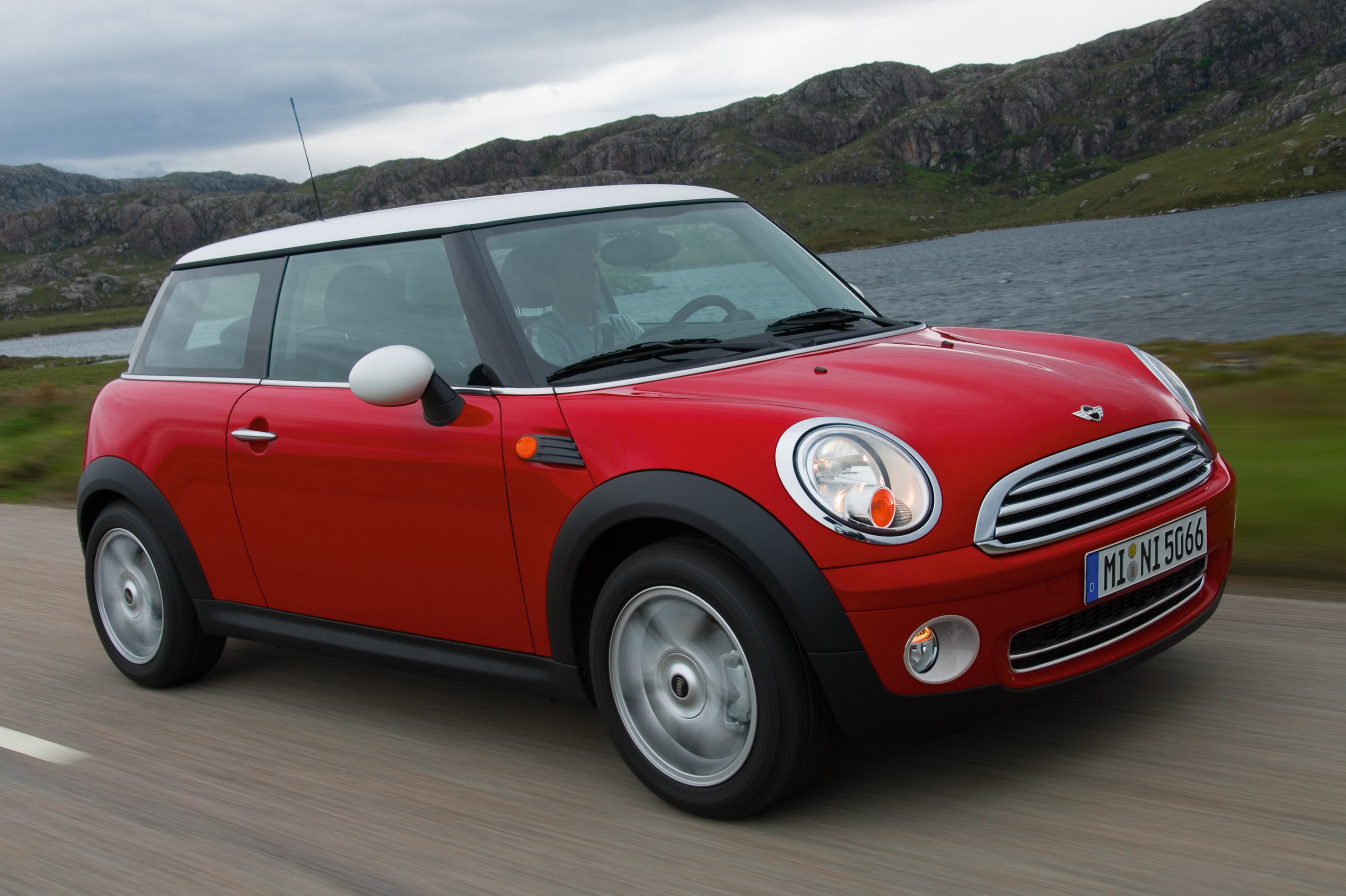A red 2007 R56 Mini Cooper driving down a road next to a hillside lake