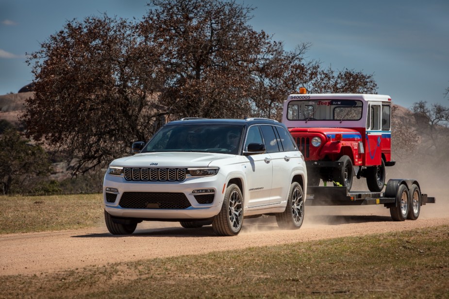 Plug-in hybrid Jeep Grand Cherokee SUV towing a classic Wrangler predecessor down a dirt road with trees in the background.