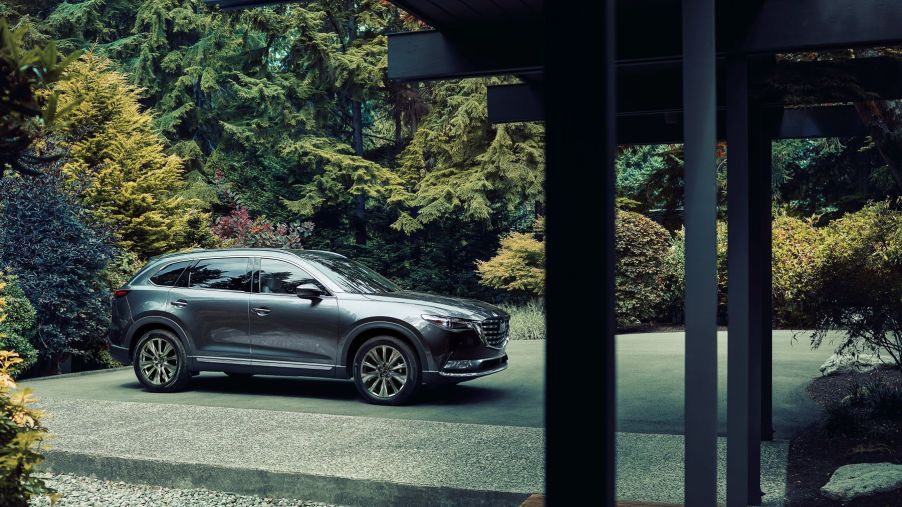 A dark gray 2022 Mazda CX-9 midsize crossover SUV parked on a gravel plaza outside a luxury home in a forest
